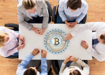 Six people sit around a white table, seen from above, with a large digital Bitcoin symbol displayed in the center of the table, suggesting a discussion or meeting about cryptocurrency. | GoCryptoBet.com