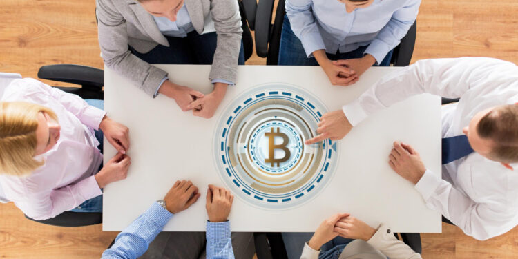 Six people sit around a white table, seen from above, with a large digital Bitcoin symbol displayed in the center of the table, suggesting a discussion or meeting about cryptocurrency. | GoCryptoBet.com