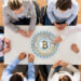 Six people sit around a white table, seen from above, with a large digital Bitcoin symbol displayed in the center of the table, suggesting a discussion or meeting about cryptocurrency. | GoCryptoBet.com