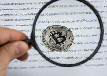 | GoCryptoBet.com A hand holds a magnifying glass over a physical Bitcoin coin placed on paper printed with binary code (ones and zeros) in rows.