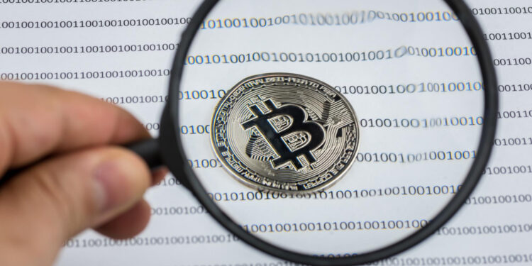 | GoCryptoBet.com A hand holds a magnifying glass over a physical Bitcoin coin placed on paper printed with binary code (ones and zeros) in rows.