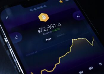 A smartphone screen displays a Bitcoin price chart with the price at $72,891.30, showing a +1.67% increase. The yellow line graph indicates recent price changes on a dark background. | GoCryptoBet.com