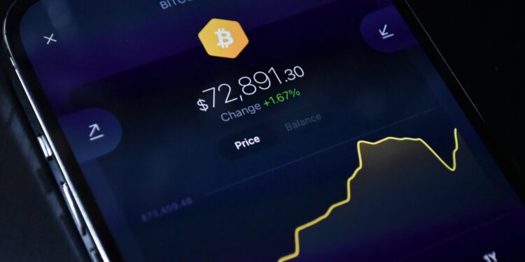 A smartphone screen displays a Bitcoin price chart with the price at $72,891.30, showing a +1.67% increase. The yellow line graph indicates recent price changes on a dark background. | GoCryptoBet.com