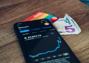 A smartphone showing a Bitcoin price chart sits on a wooden surface next to a rainbow-colored card and a folded five-dollar bill. | GoCryptoBet.com