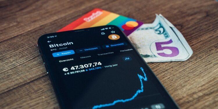 A smartphone showing a Bitcoin price chart sits on a wooden surface next to a rainbow-colored card and a folded five-dollar bill. | GoCryptoBet.com