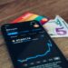 A smartphone showing a Bitcoin price chart sits on a wooden surface next to a rainbow-colored card and a folded five-dollar bill. | GoCryptoBet.com